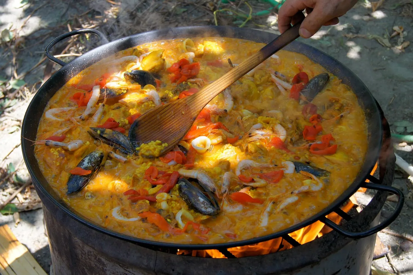 A wide paella pan over a fire, with mussels, squid and red peppers in the rice and a wooden paddle
