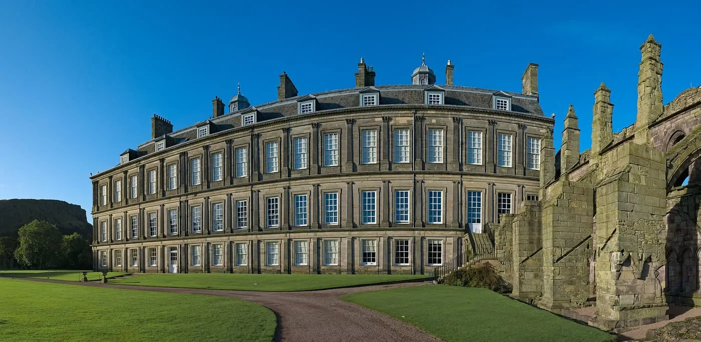 The curved stone front of the Palace of Holyroodhouse above a lawn, with the carved stonework of the abbey ruins at the right edge