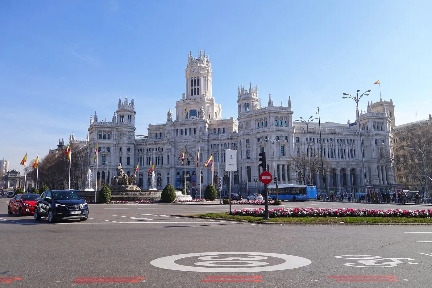 The white stone Palacio de Cibeles with its clock tower and flagpoles, seen across the Plaza de Cibeles under a blue sky