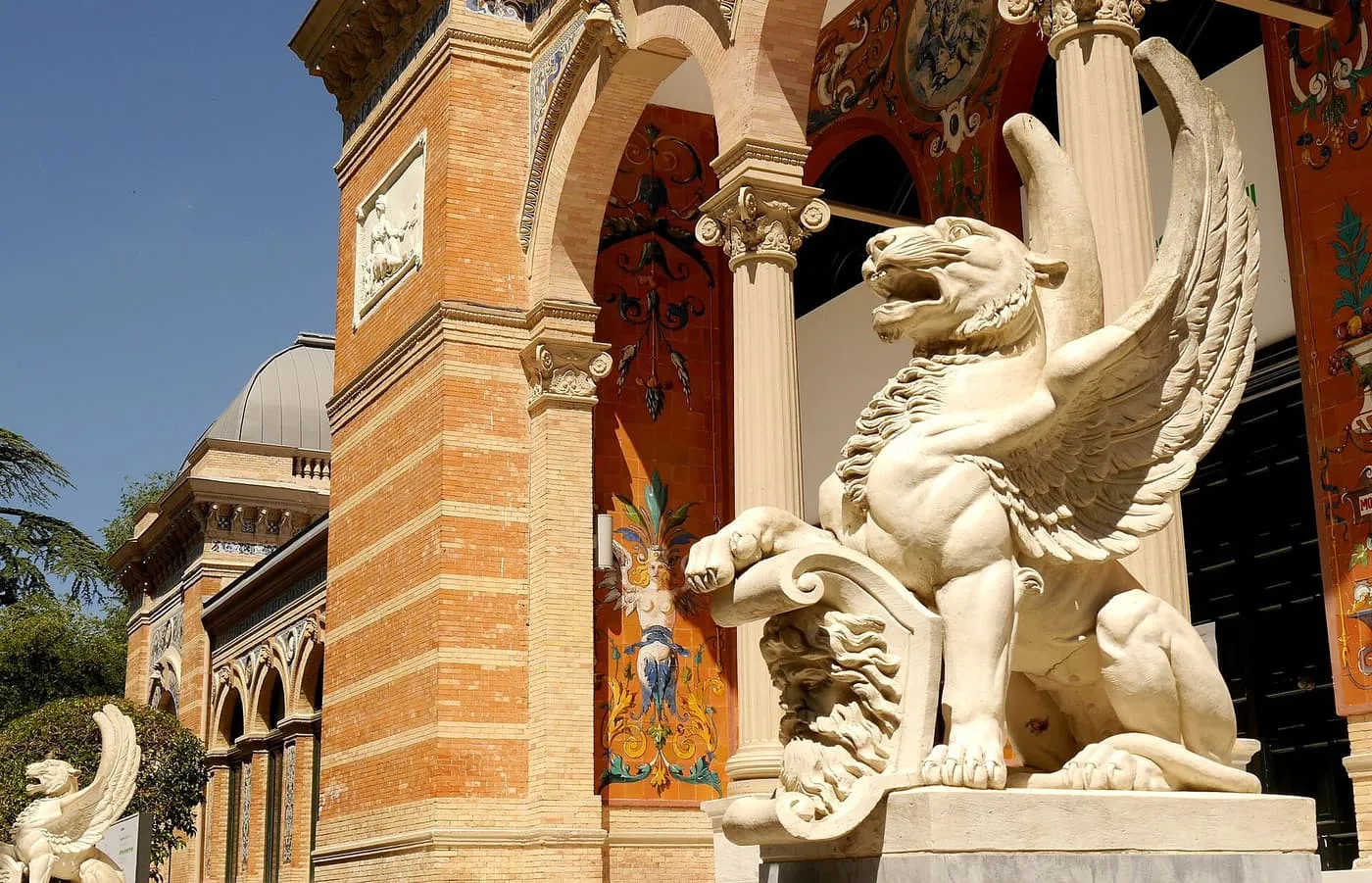 A white stone winged lion beside the entrance of the Palacio de Velázquez, with its brick walls and painted tiles behind