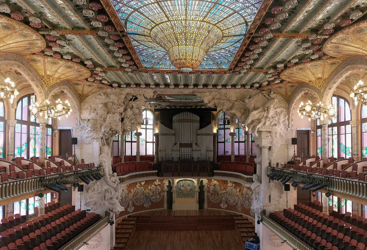 The concert hall of the Palau de la Música Catalana seen from the gallery, with the stained-glass skylight above rows of red seats and the organ over the stage