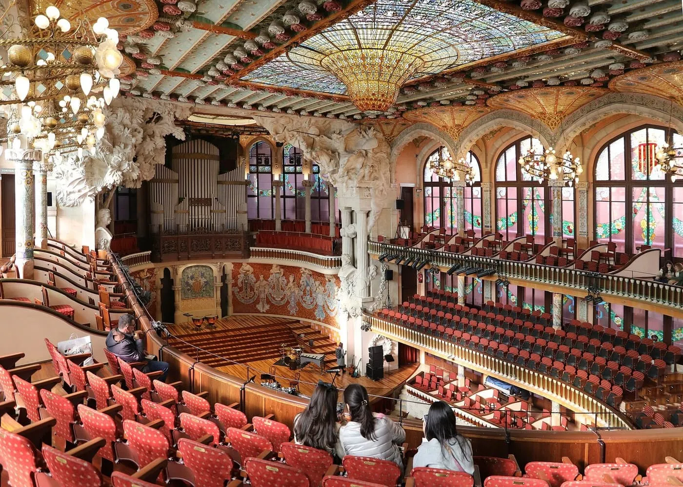 The concert hall of the Palau de la Música Catalana seen from the gallery: red seats, a stained-glass skylight and ornate mosaic-covered balconies