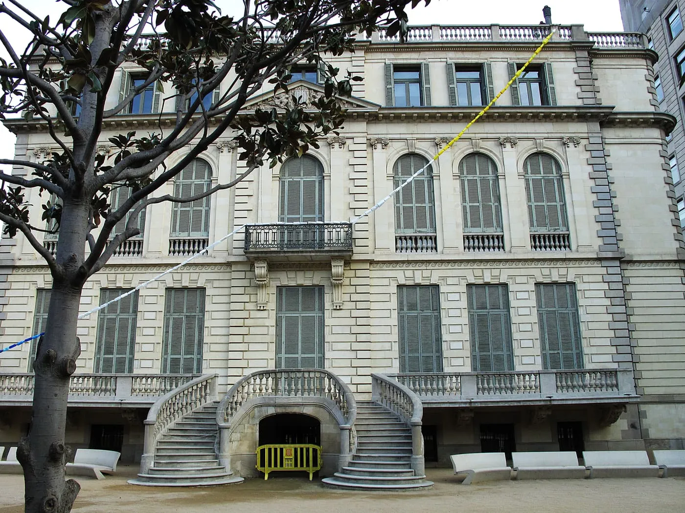 The garden front of Palau Robert, a stone neoclassical palace with a double curved staircase, tall arched windows and a magnolia tree in front