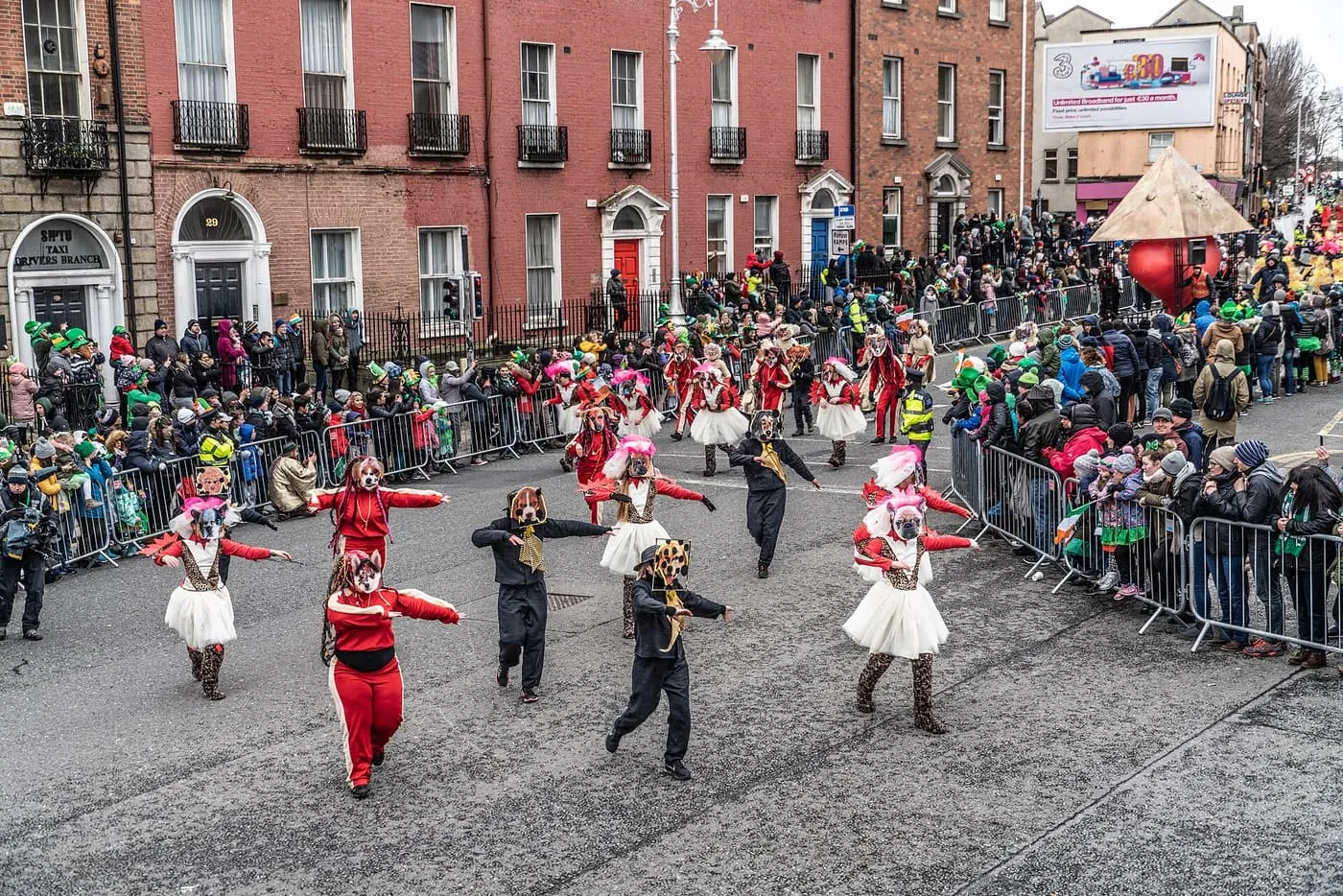 Performers in red and white costumes and animal masks dancing in the street in front of crowds, with a float behind them