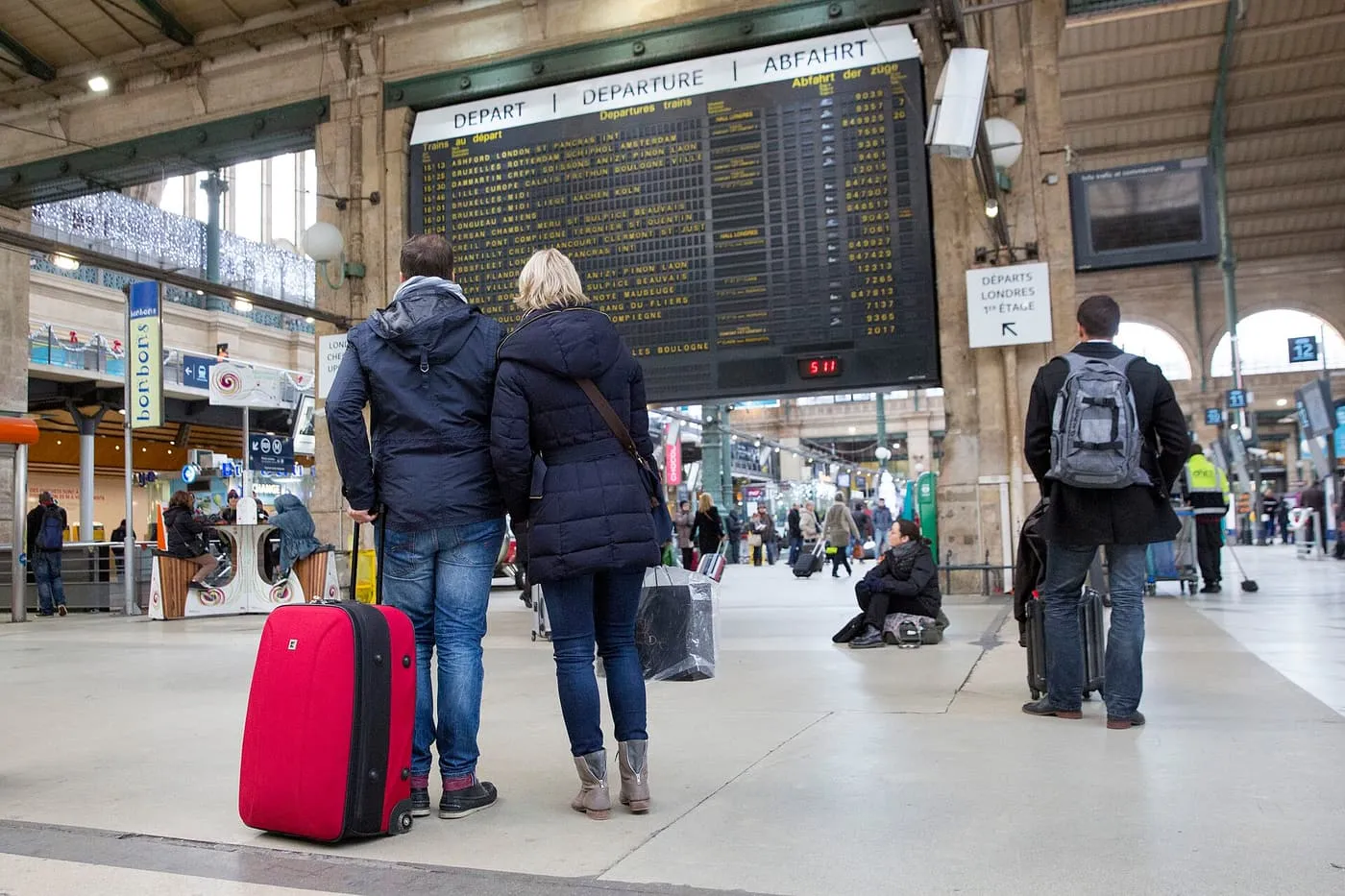 Two travellers with a red suitcase facing the departures board at Gare du Nord in Paris, which lists trains to London and Amsterdam