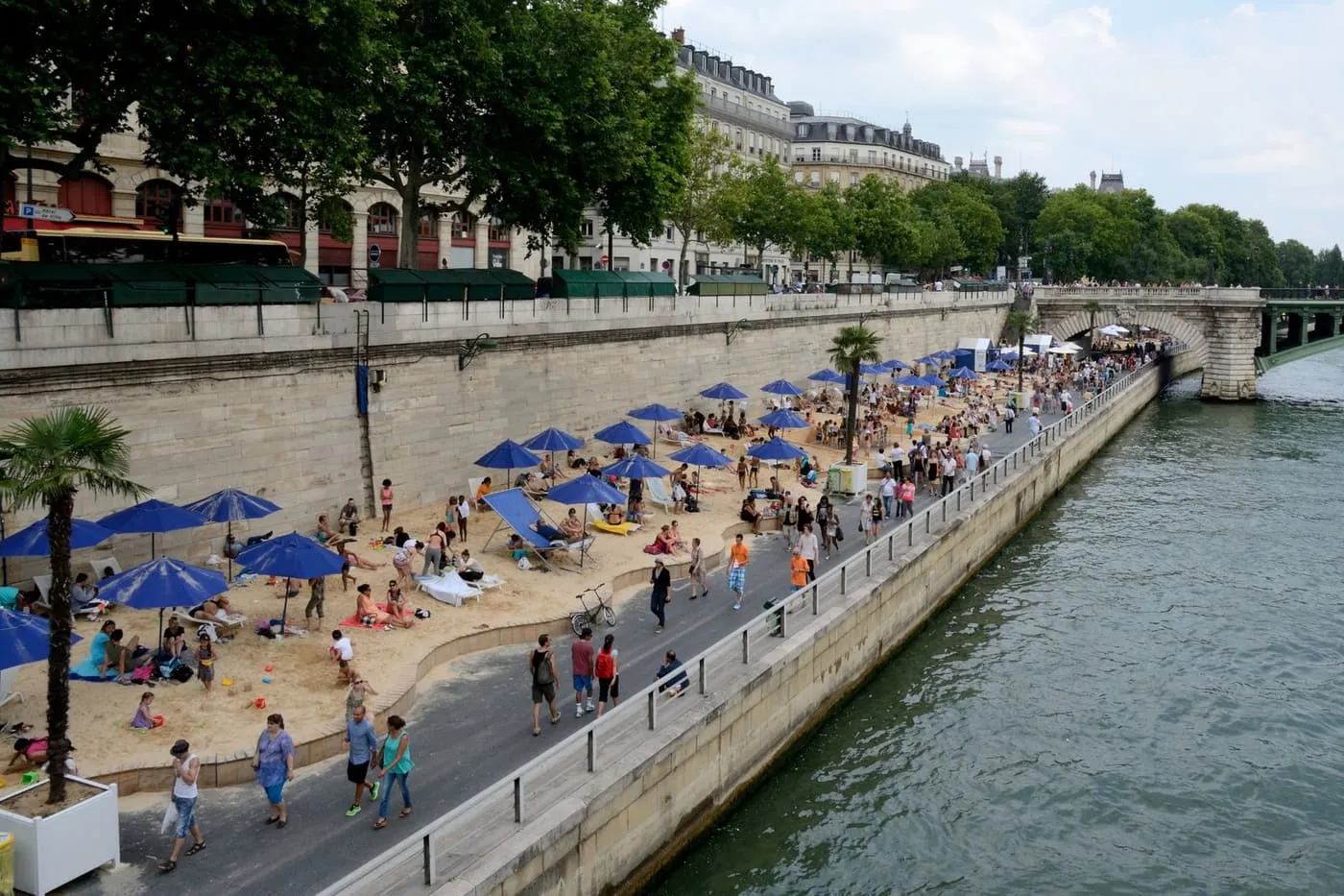 Sand, blue parasols and deckchairs of the Paris Plages beach beside the Seine, with people walking along the riverside road and a bridge beyond