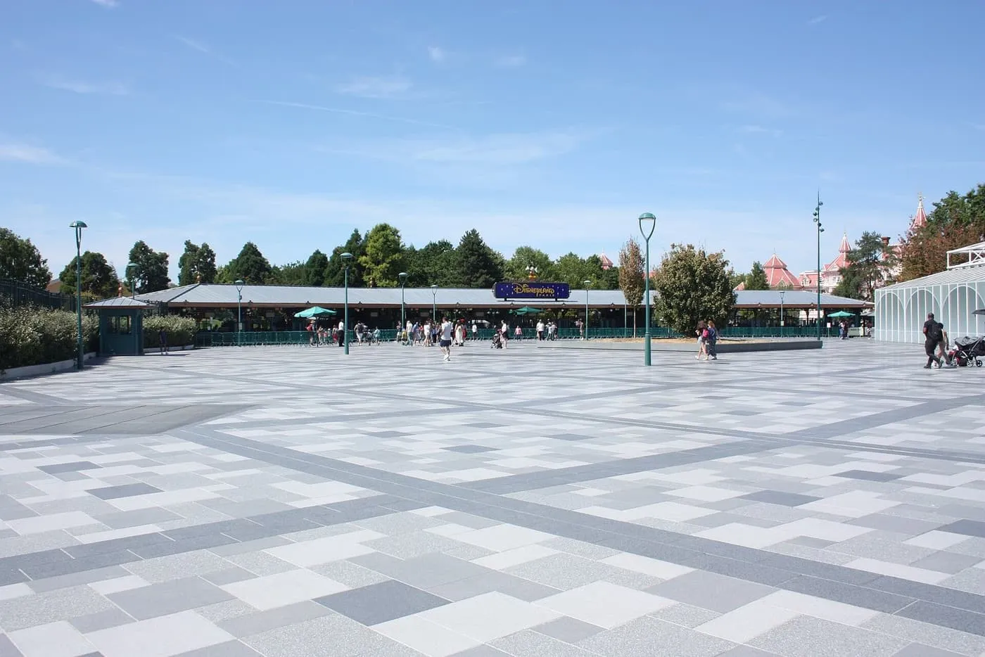 The paved plaza in front of the ticket gates of Disneyland Park, with visitors walking towards the entrance gates under a long low canopy