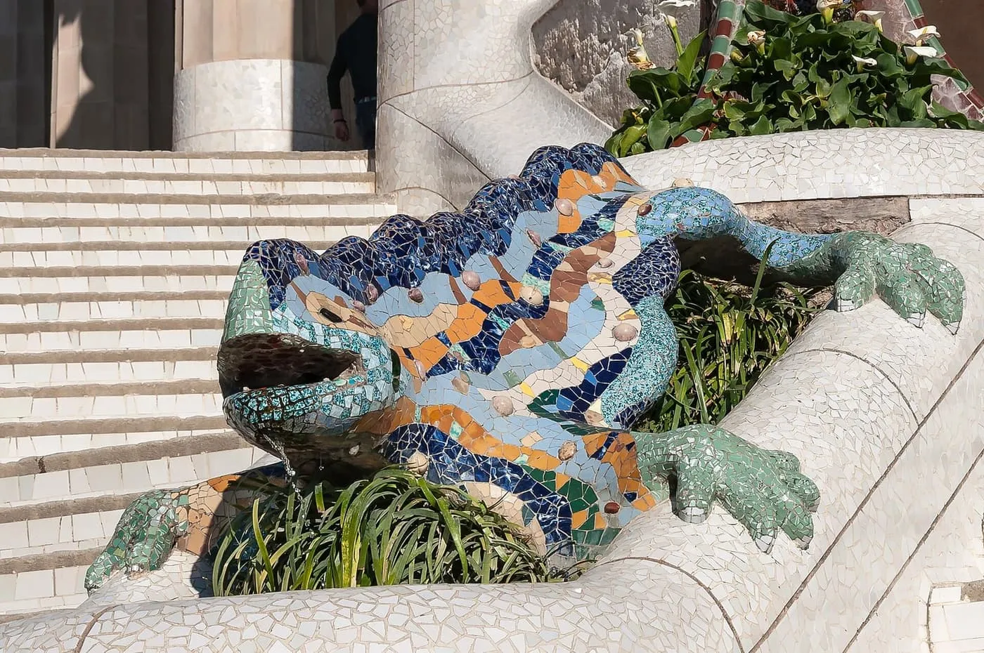 A mosaic salamander in blue, orange and green broken tiles, water running from its mouth, on the white steps of Park Güell