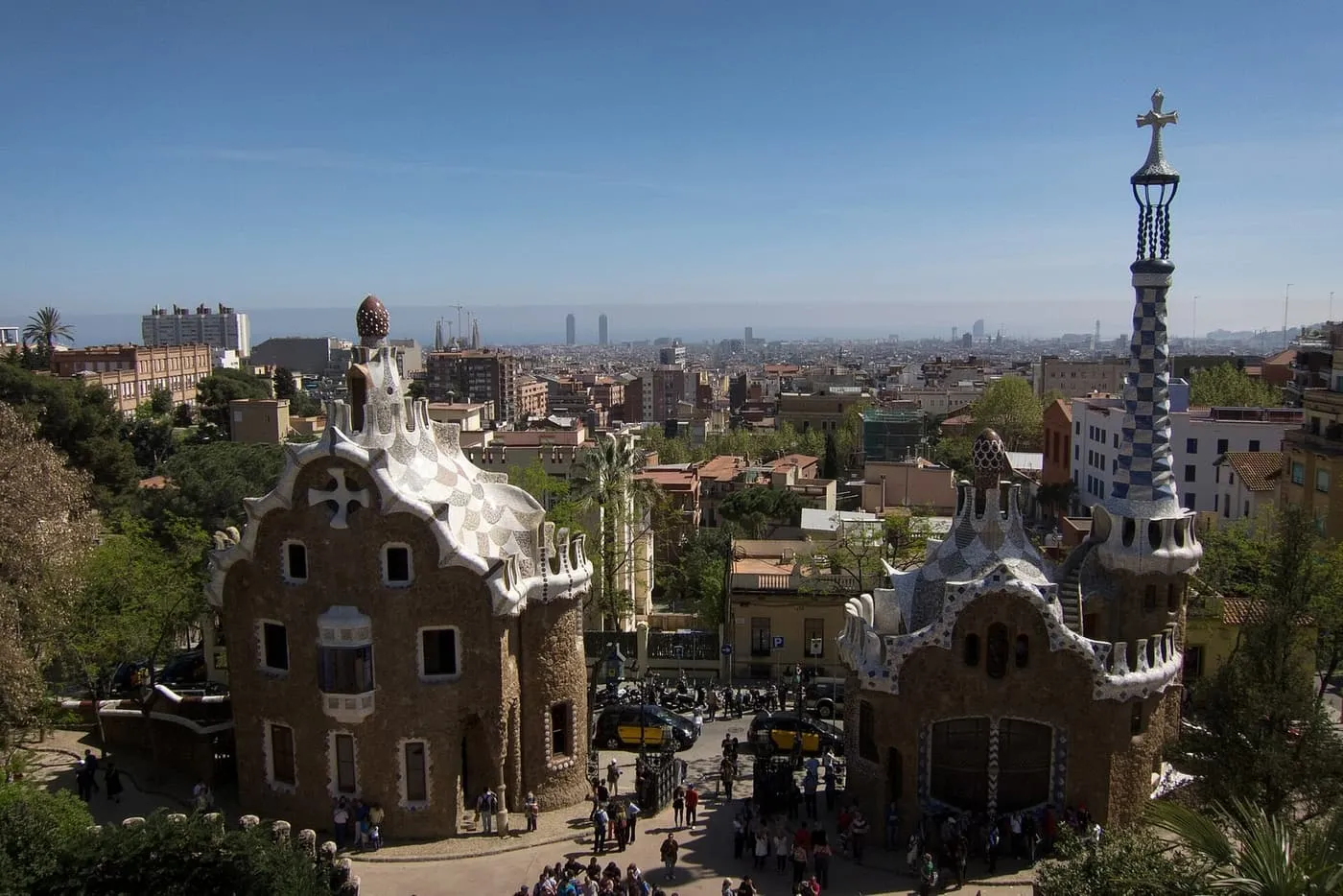 Gaudí's two gatehouse pavilions, with white mosaic roofs and a chequered blue tower topped by a cross, and the city stretching to the sea behind
