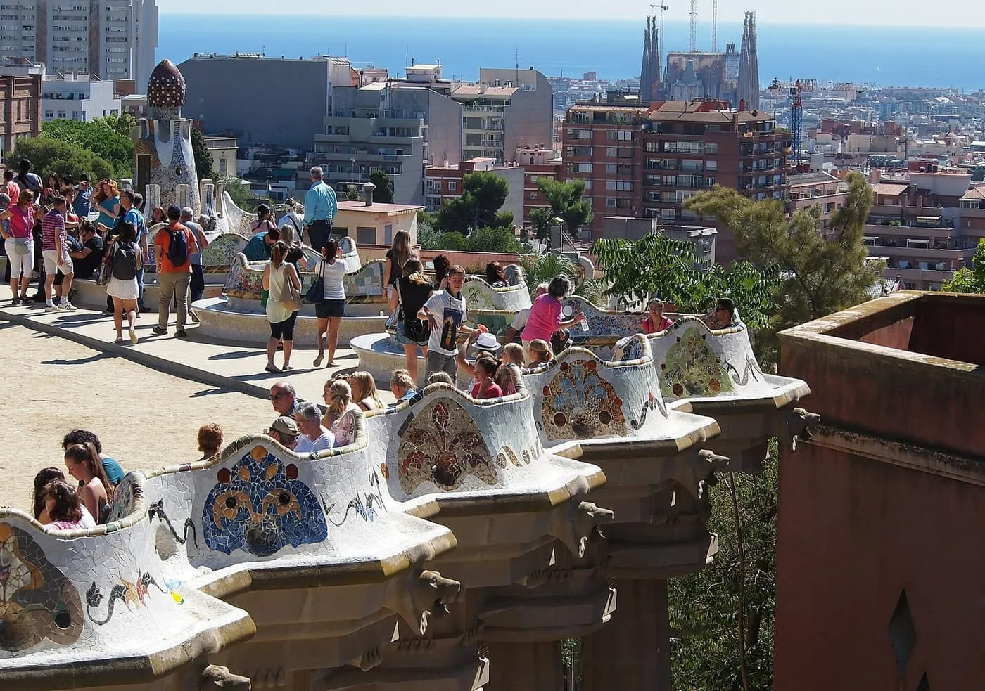 Visitors along the curving mosaic bench of Park Güell's Nature Square, with the Sagrada Família and the sea beyond the rooftops