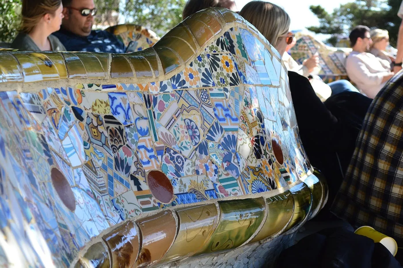 The curving mosaic serpentine bench of Park Güell in broken blue, white and yellow tile, with visitors seated along it