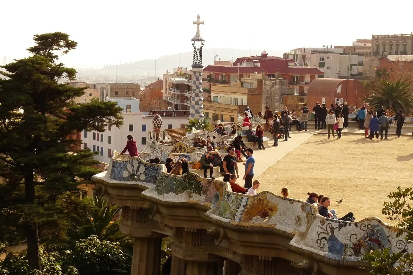 Visitors on the mosaic-tiled serpentine bench round Park Güell's terrace, with a chequered tower topped by a cross behind