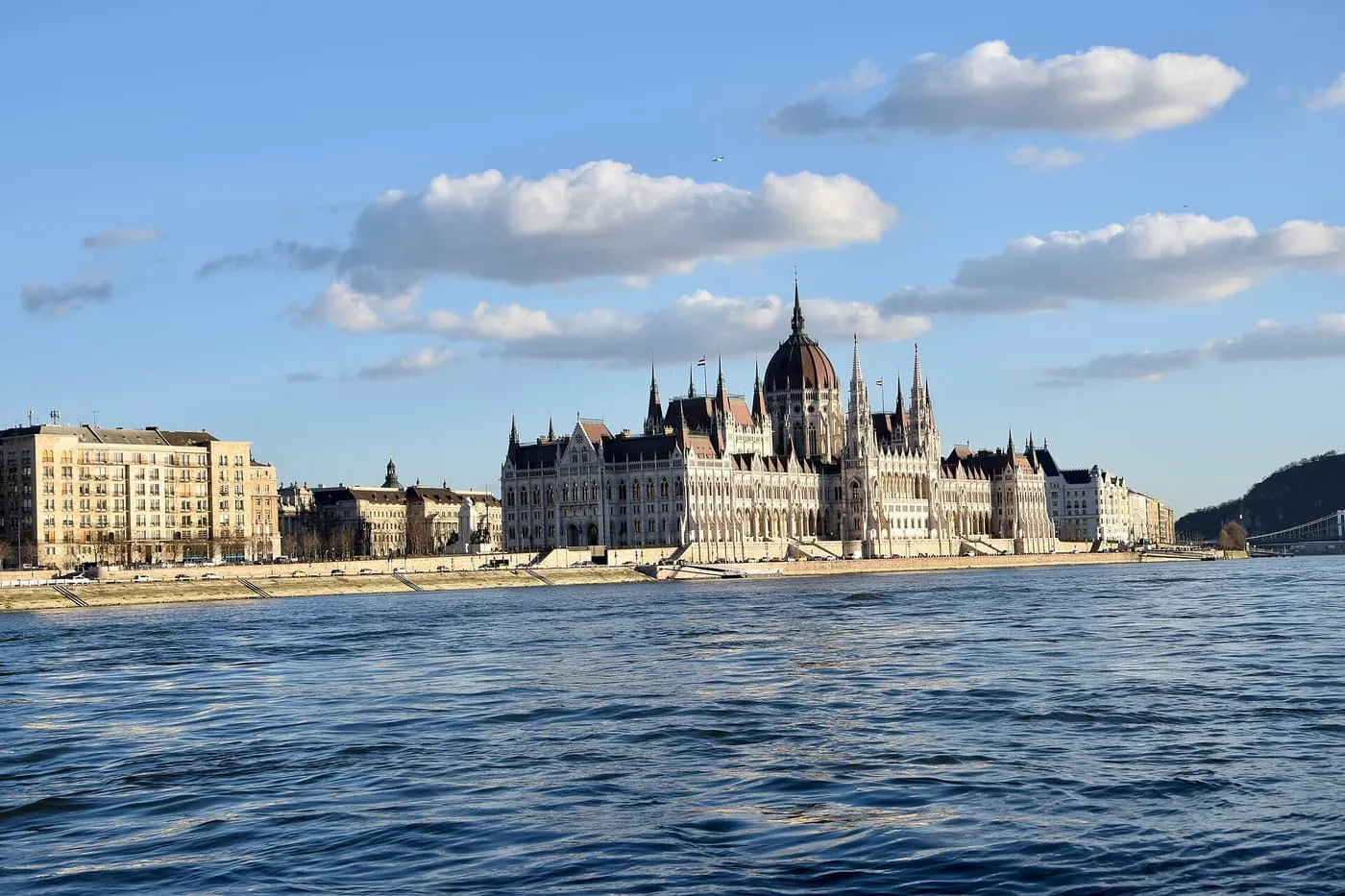 The Hungarian Parliament Building on the Pest bank of the Danube, seen from the water, with a wooded hill and a bridge beyond on the right