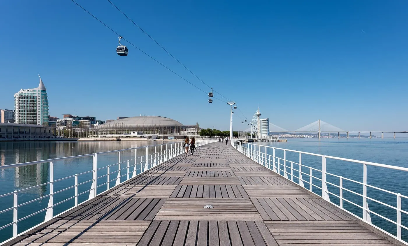 A wooden boardwalk beside the Tagus with white railings, a cable car overhead and the Vasco da Gama Bridge in the distance