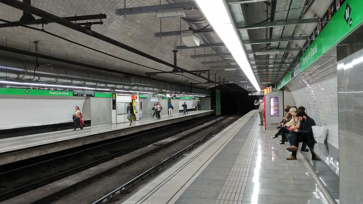 The L3 platforms at Passeig de Gràcia metro station, with green station signs, a few passengers walking and others seated by the wall