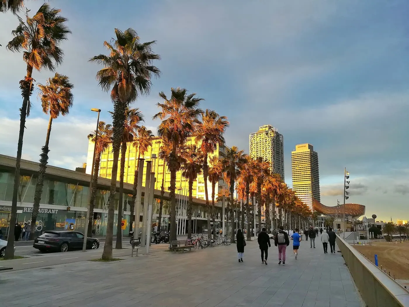 Palms and walkers on the Passeig Marítim at dusk, with two tall towers, Hotel Arts and the Torre Mapfre, rising over the Port Olímpic