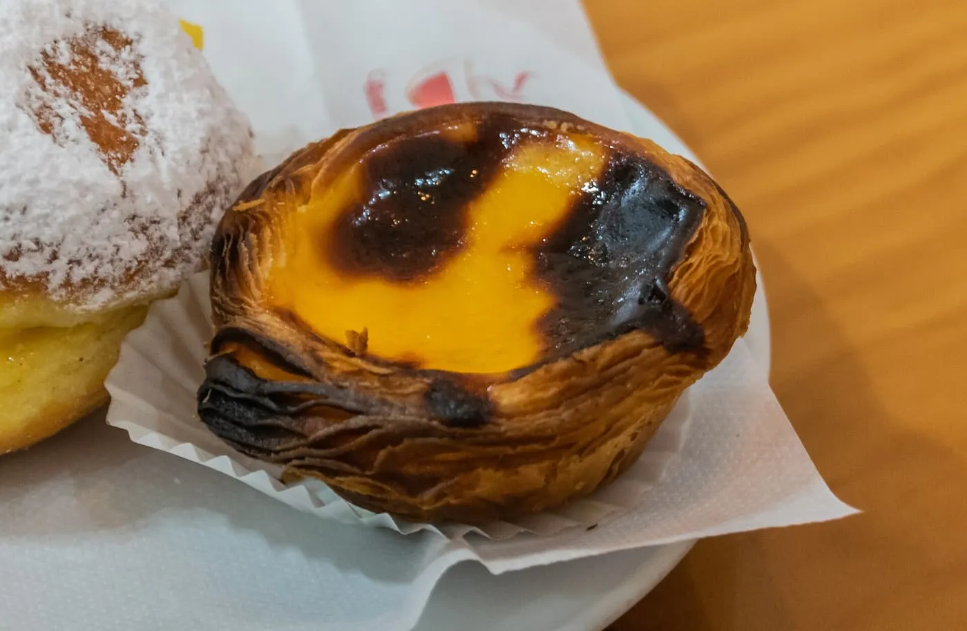 A pastel de nata with a blistered, caramelised custard top in a paper case on a white plate, beside a sugar-dusted pastry