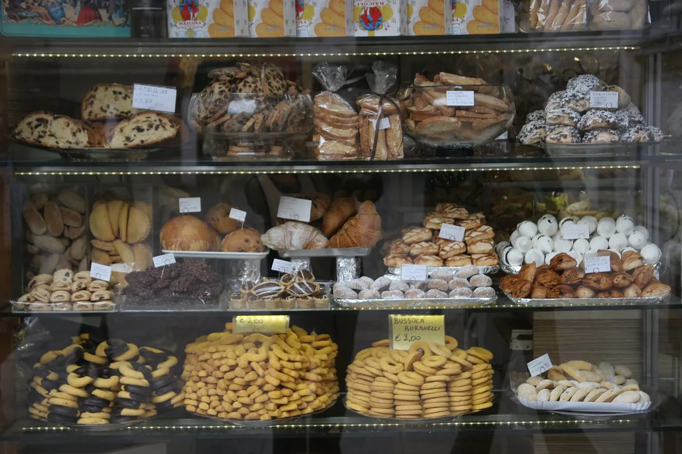 A glass display of filled pastries, croissants and trays of ring-shaped biscuits with handwritten price labels