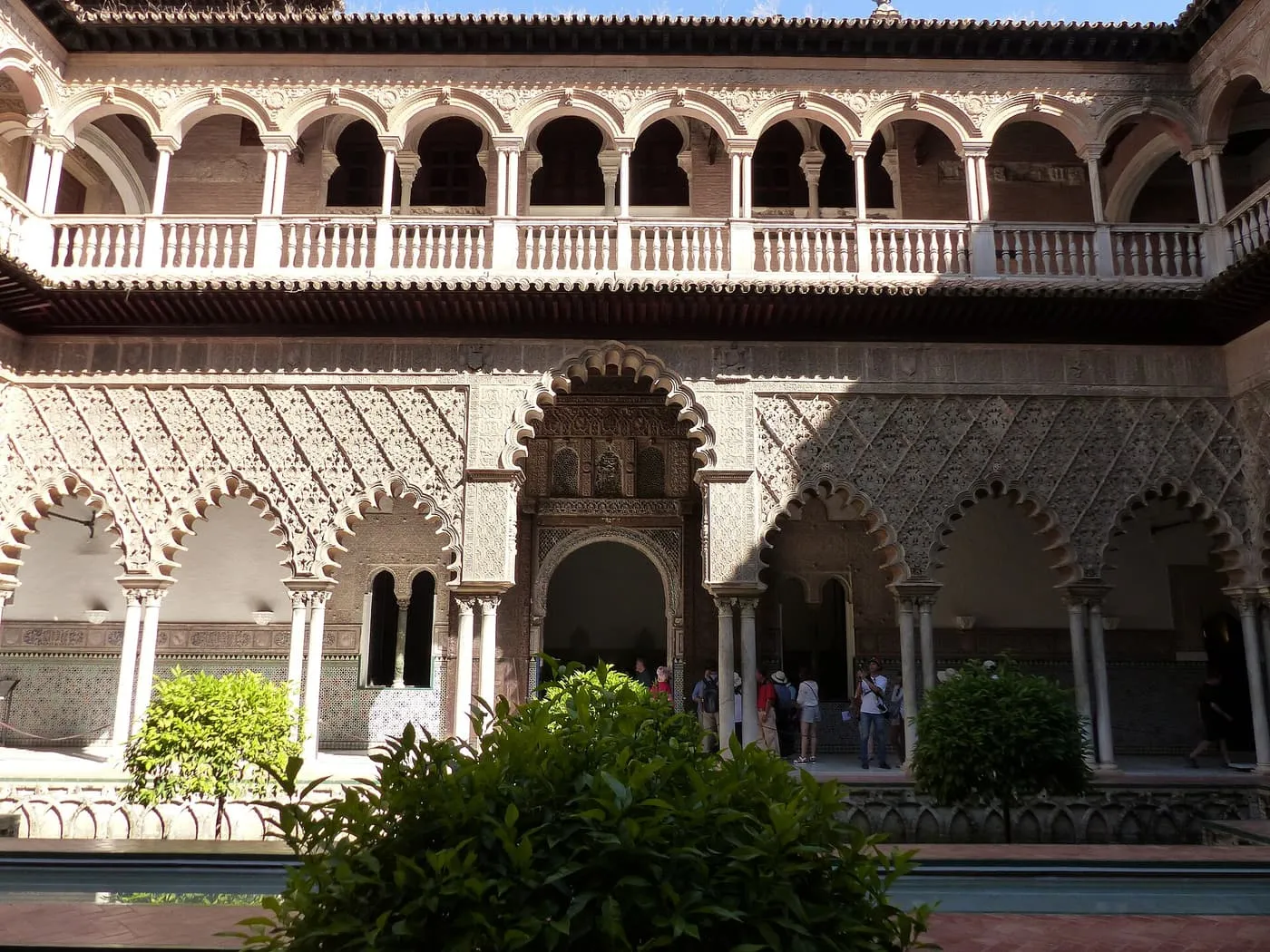 The arcaded Patio de las Doncellas in the Real Alcázar, with carved plasterwork arches below a two-storey gallery and visitors at the far side