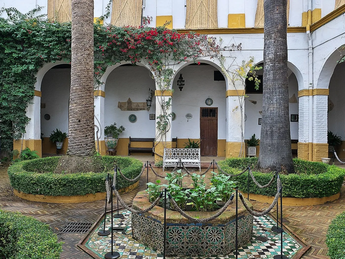 A tiled fountain between two palm trunks in a whitewashed courtyard hung with bougainvillea