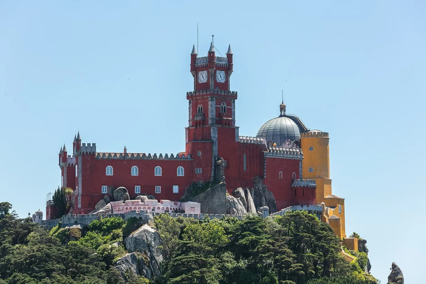 The red clock tower and yellow dome of Pena Palace on its hilltop above the trees, against a blue sky
