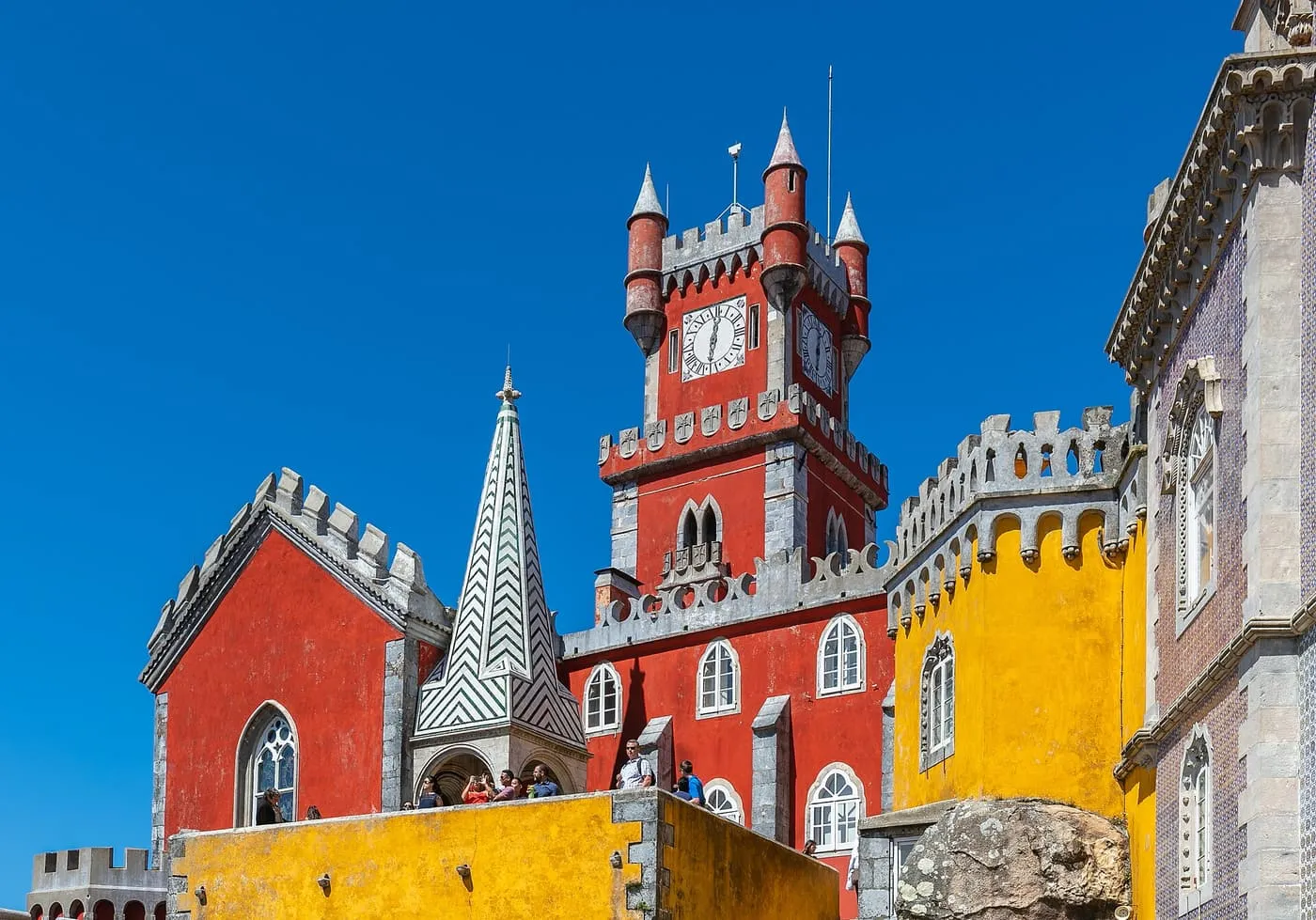 Pena Palace's red clock tower beside a yellow wall and a green-and-white striped spire, against a deep blue sky