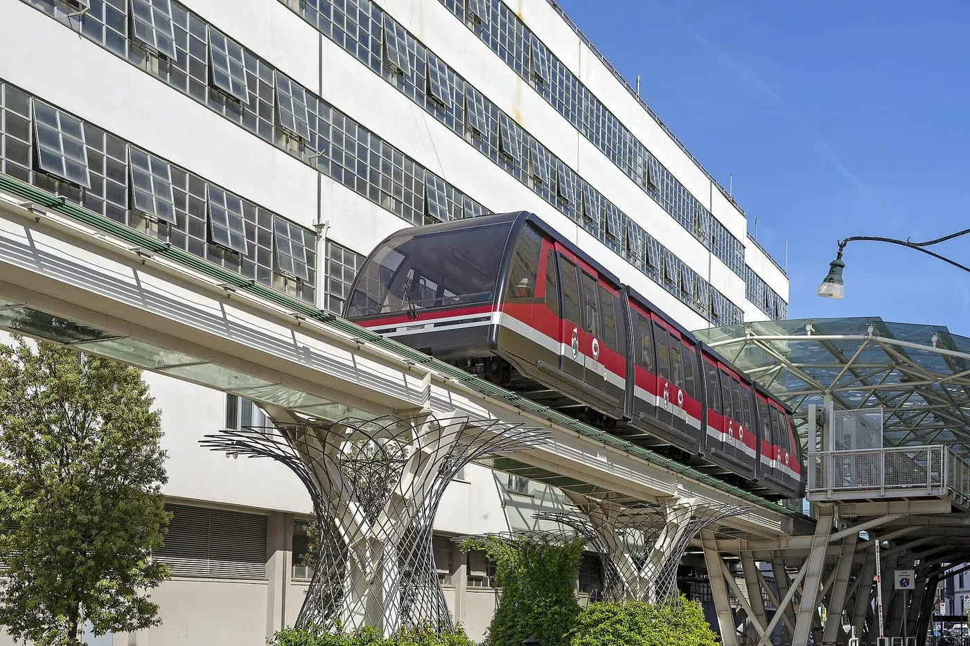 A red and grey People Mover train on its raised track beside a long white building