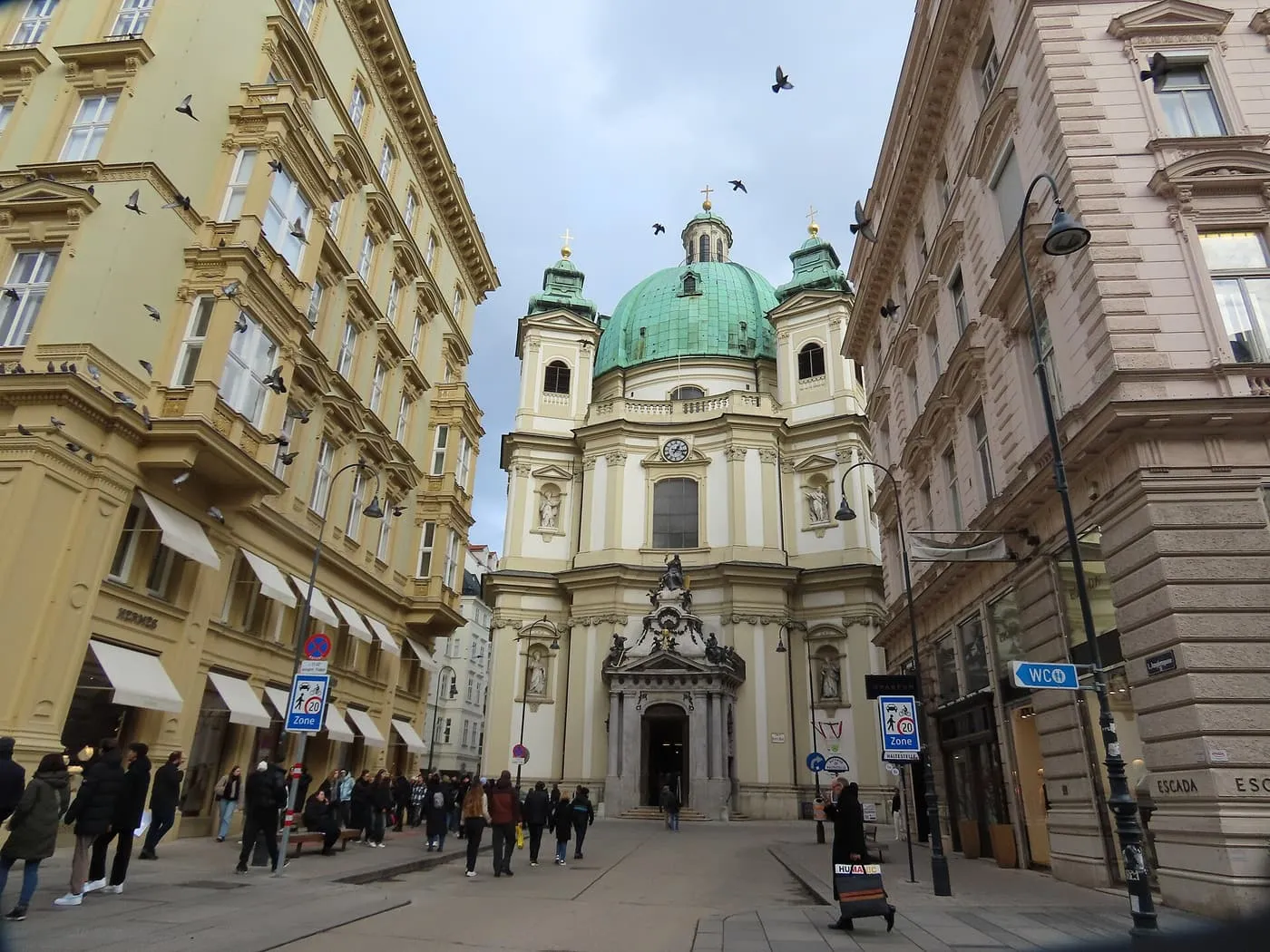 The green-domed Peterskirche at the end of a street of tall cream and pink buildings on Petersplatz, with pigeons overhead