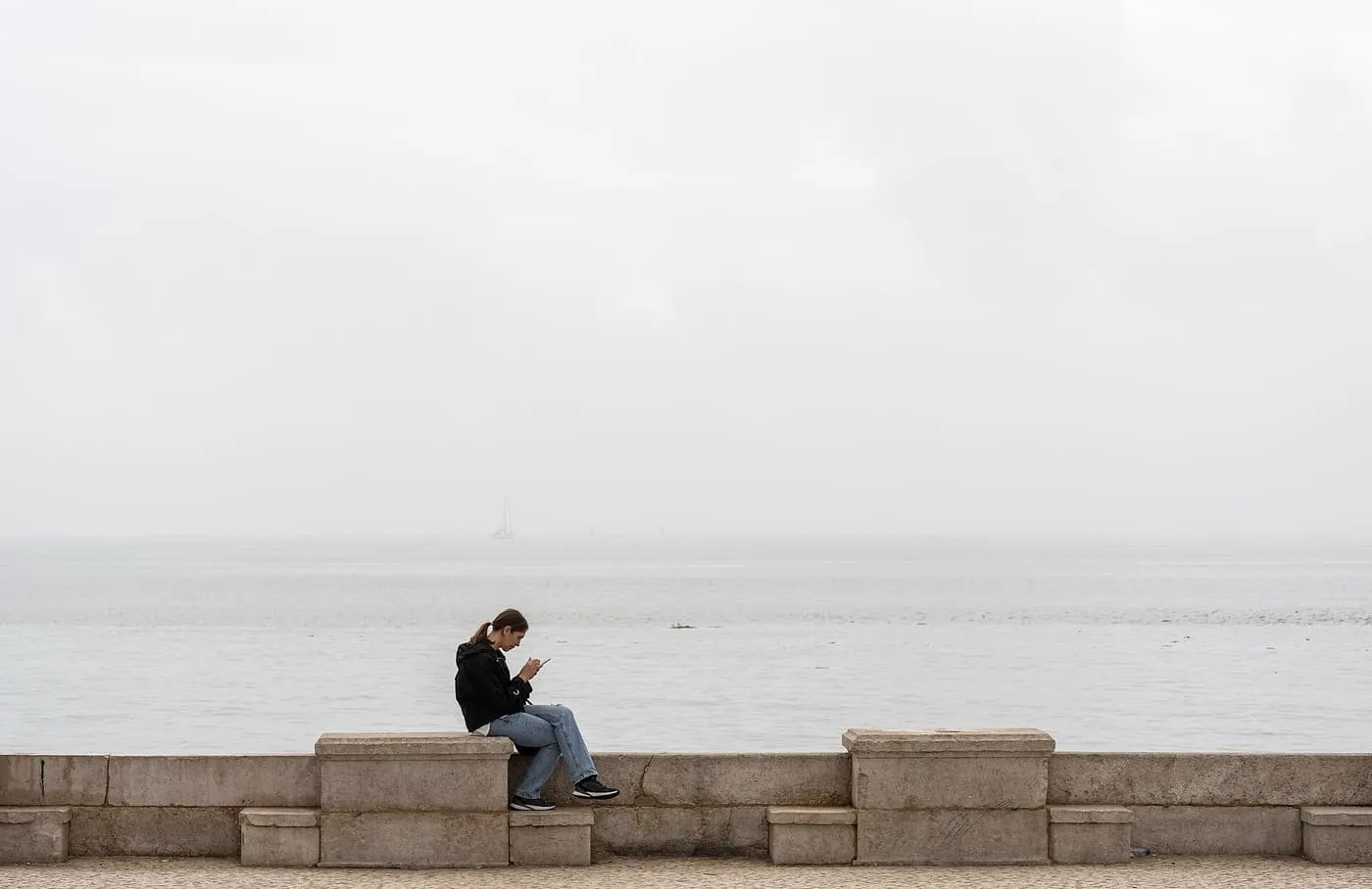 A woman sitting on a stone quay wall using her phone, with the hazy Tagus and a sailing boat behind her