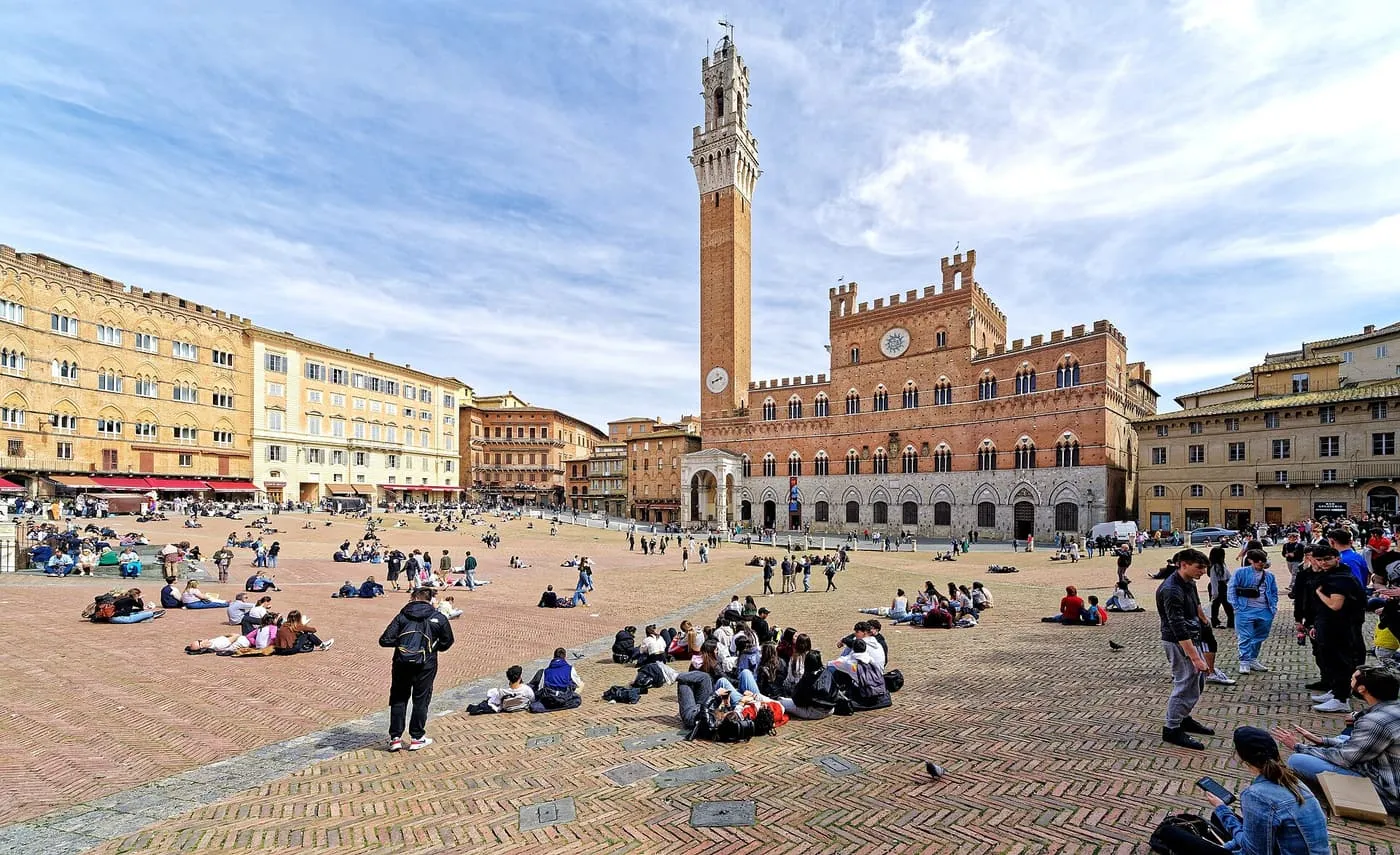 The Torre del Mangia and the Palazzo Pubblico at the edge of the sloping brick Piazza del Campo, with people sitting on the paving