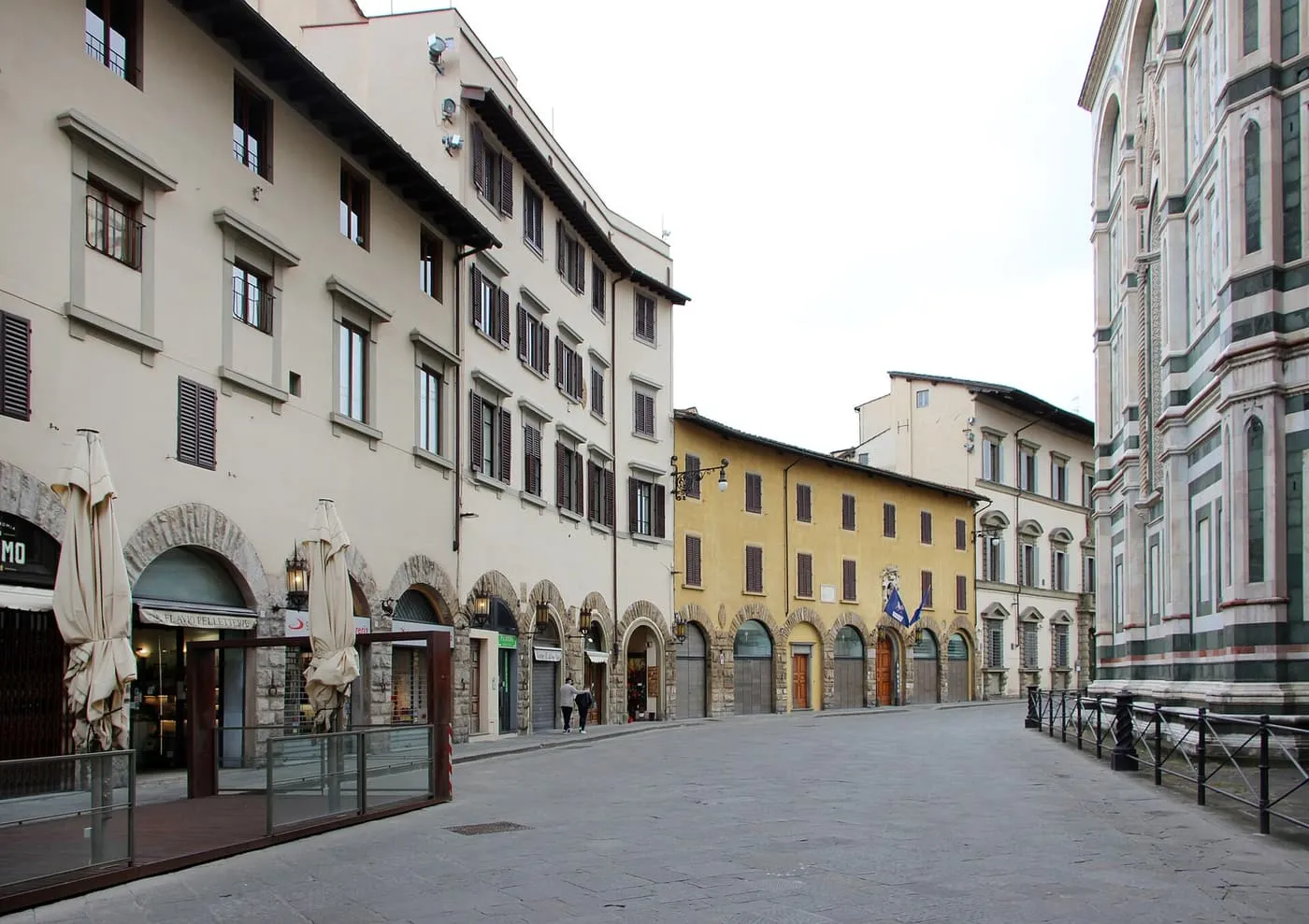 A street of pale shuttered buildings with arcaded shopfronts running towards the marble flank of the cathedral at right
