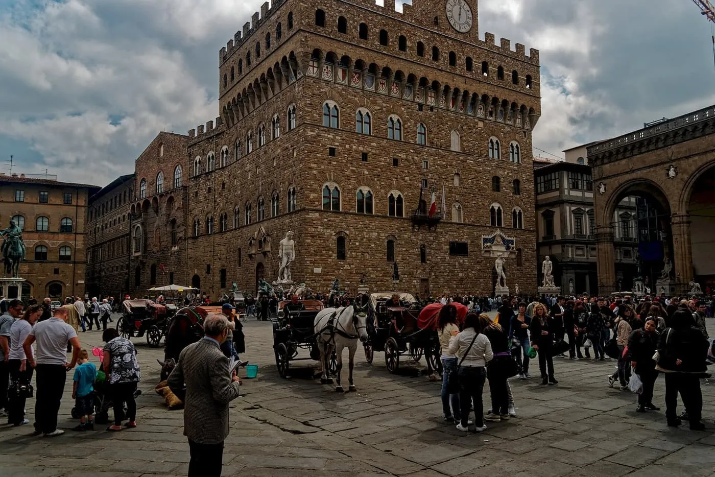 Palazzo Vecchio's tower and crenellated walls above horse-drawn carriages and crowds in Piazza della Signoria