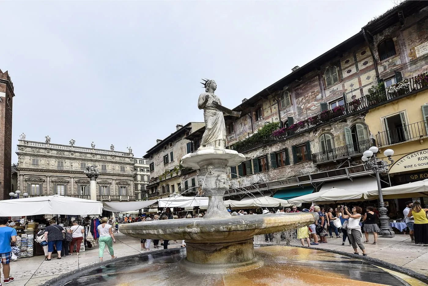 Piazza delle Erbe in Verona: a fountain topped by a statue, market stalls and café umbrellas below frescoed palace façades