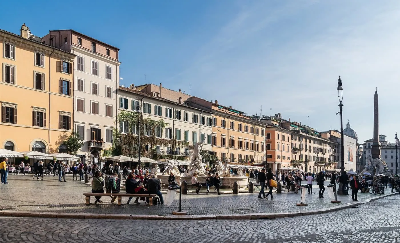 Piazza Navona, with a fountain at the centre, the obelisk of the Fountain of the Four Rivers at the right, and restaurant awnings under the ochre buildings