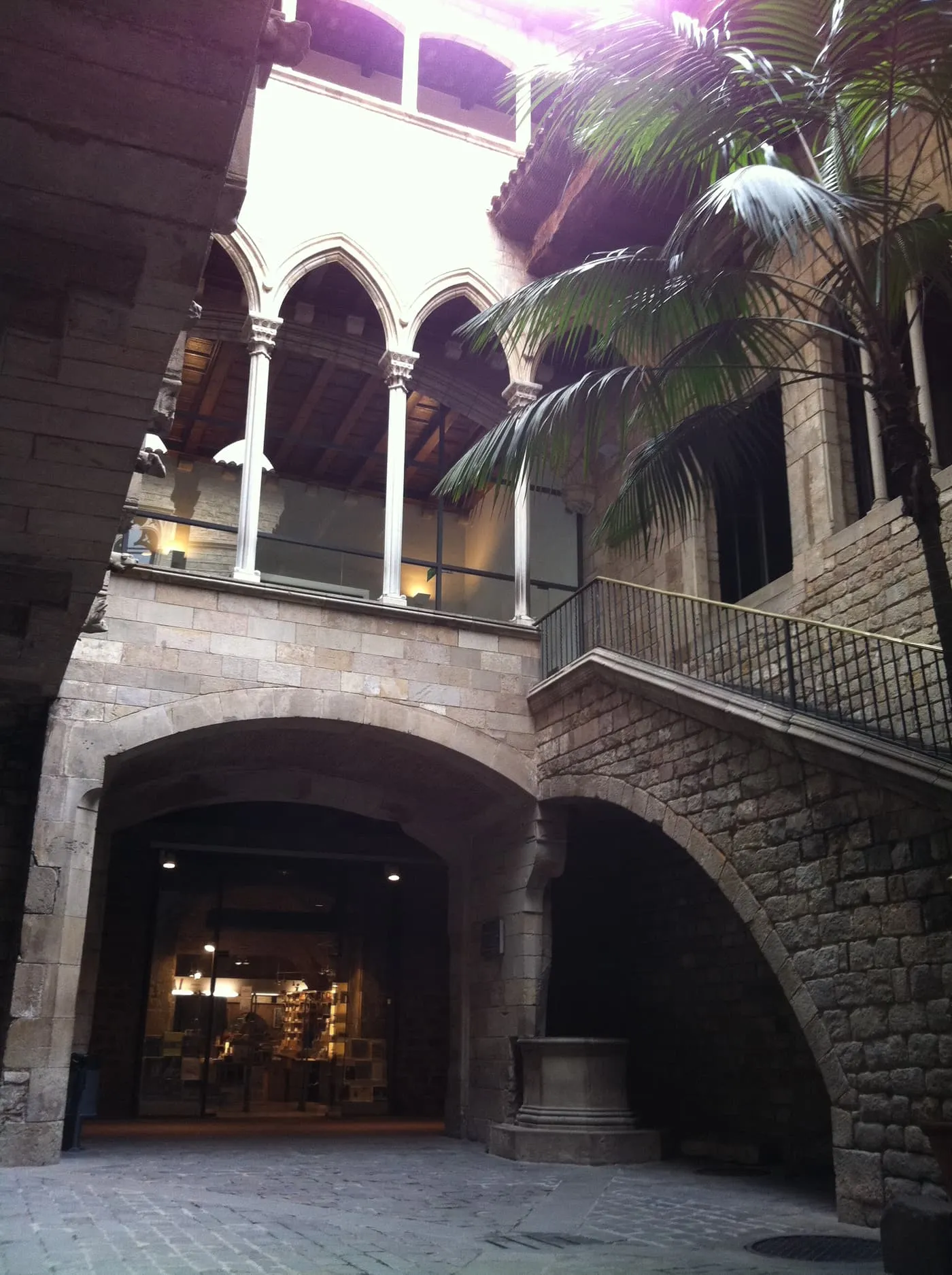 A courtyard of the Palau Aguilar, with a Gothic arcade on the upper floor, an open stone staircase and a palm tree
