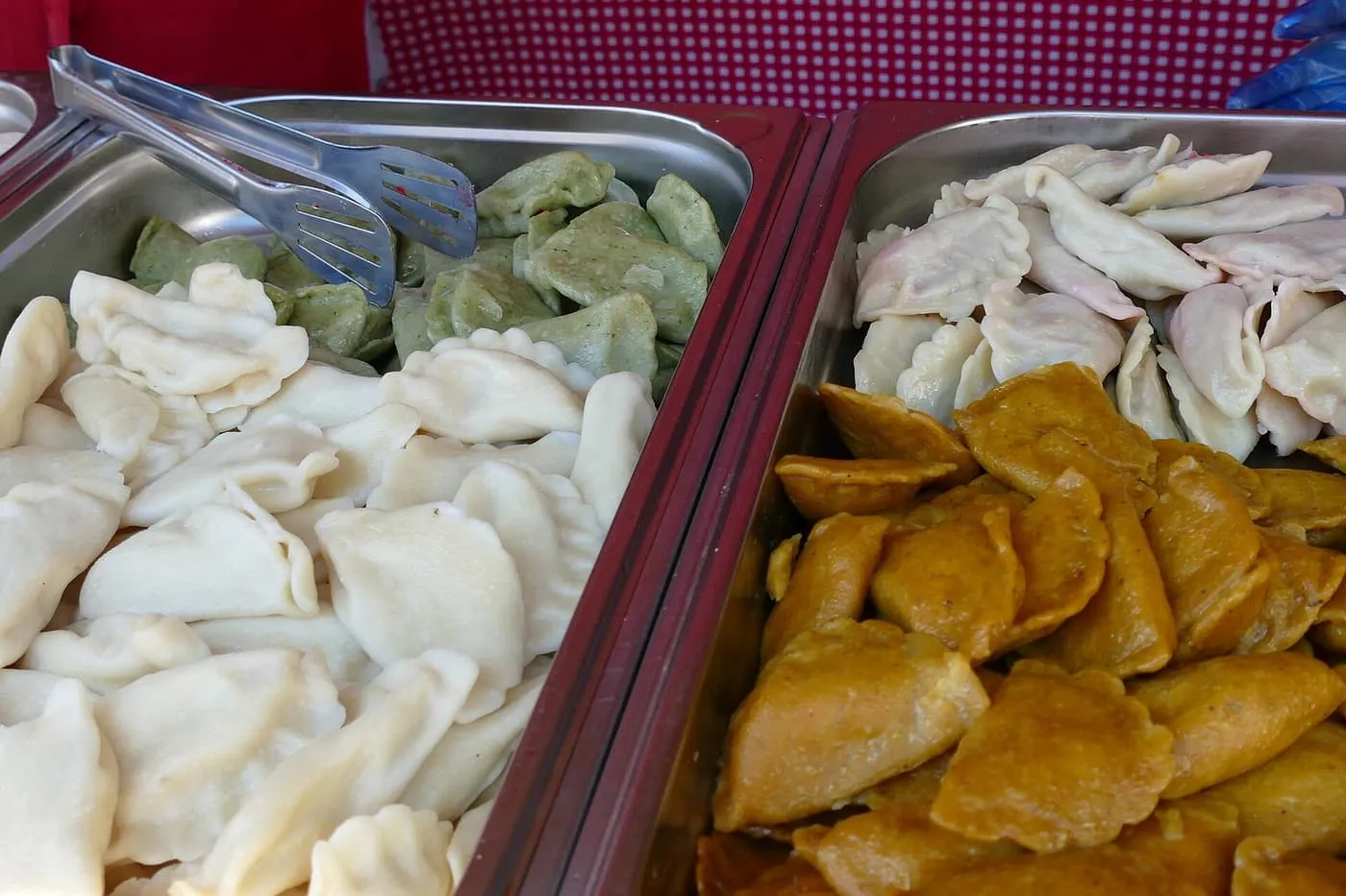 Steel trays of boiled pierogi, some plain, some green, pink and orange, with serving tongs