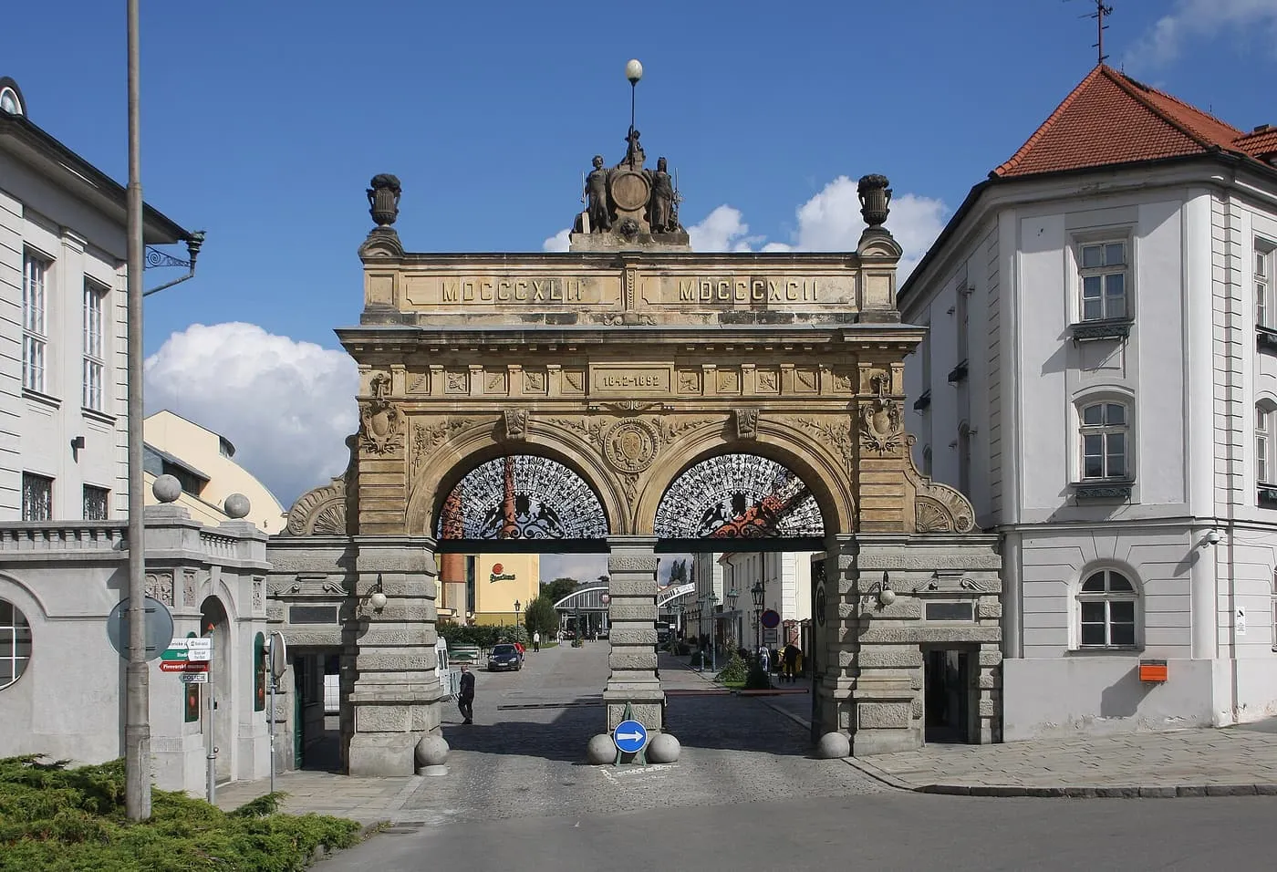 The twin-arched sandstone gate of the Pilsner Urquell brewery in Plzeň, with the dates 1842 and 1892 and wrought-iron grilles in both arches, under a blue sky
