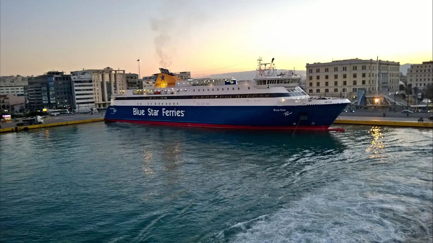 A blue and white Blue Star Ferries ship alongside the quay at Piraeus port, with the port's buildings behind in evening light