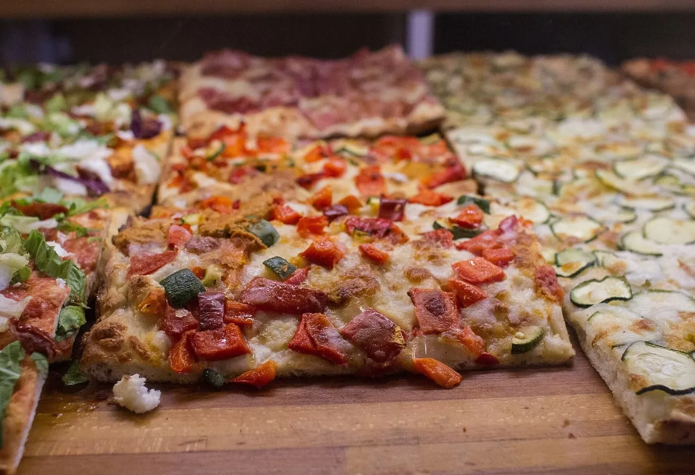 Rectangular slices of pizza al taglio topped with roasted peppers and courgette in a glass counter