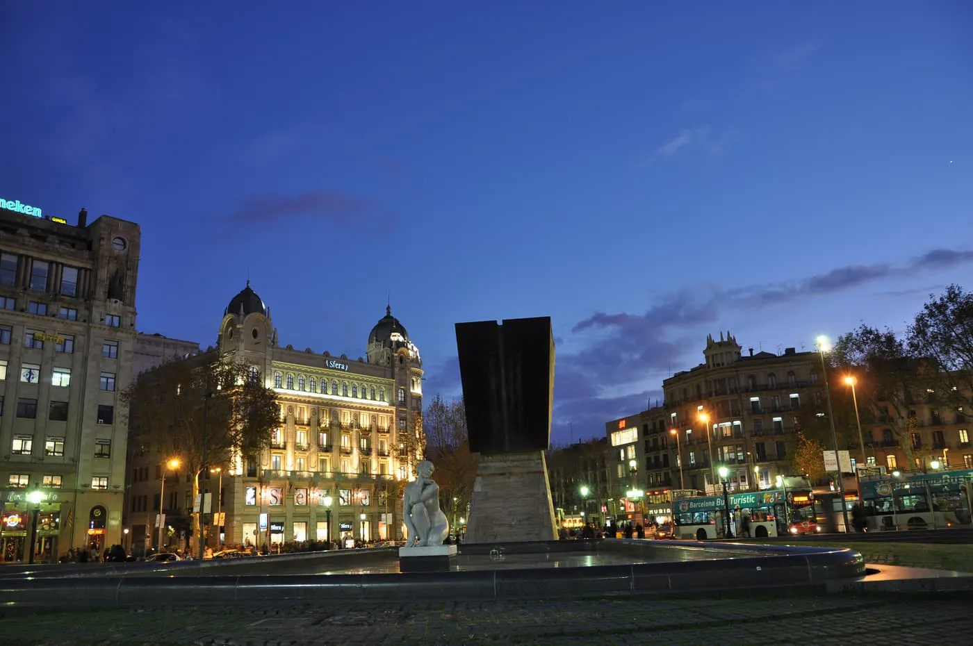 Plaça de Catalunya at dusk, with the dark monument to Francesc Macià and a domed corner building lit up behind it