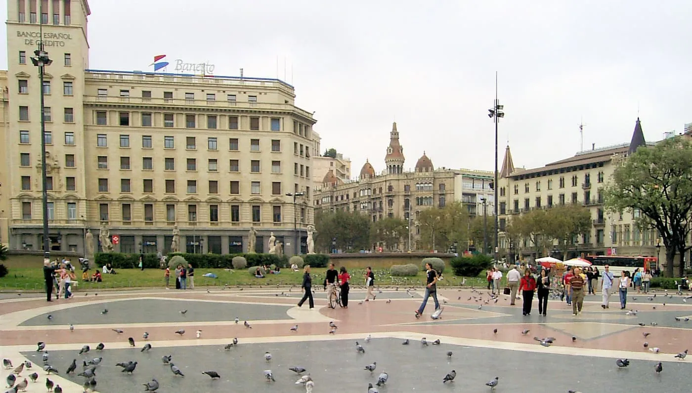 Plaça de Catalunya, a wide paved square with a pattern of pale and dark paving, pigeons in the foreground and tall stone buildings behind