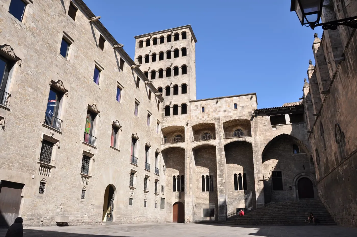 Plaça del Rei, a stone square closed in by the Gothic palace, with a tall watchtower and arcaded galleries under a blue sky