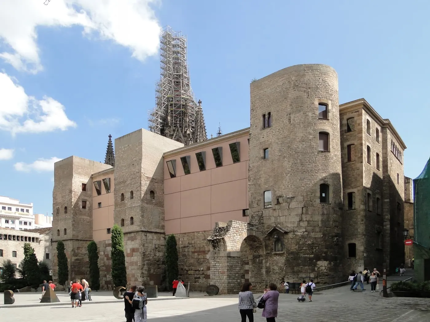Two round Roman towers and a stretch of old wall on Plaça Nova, with scaffolding on the cathedral's spire behind and a few people crossing the square
