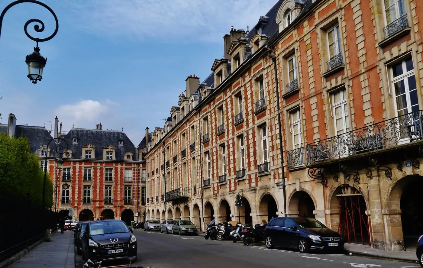 Red-brick and stone houses above an arcade on the Place des Vosges, with parked cars and scooters below and a wrought-iron lamp at the top left