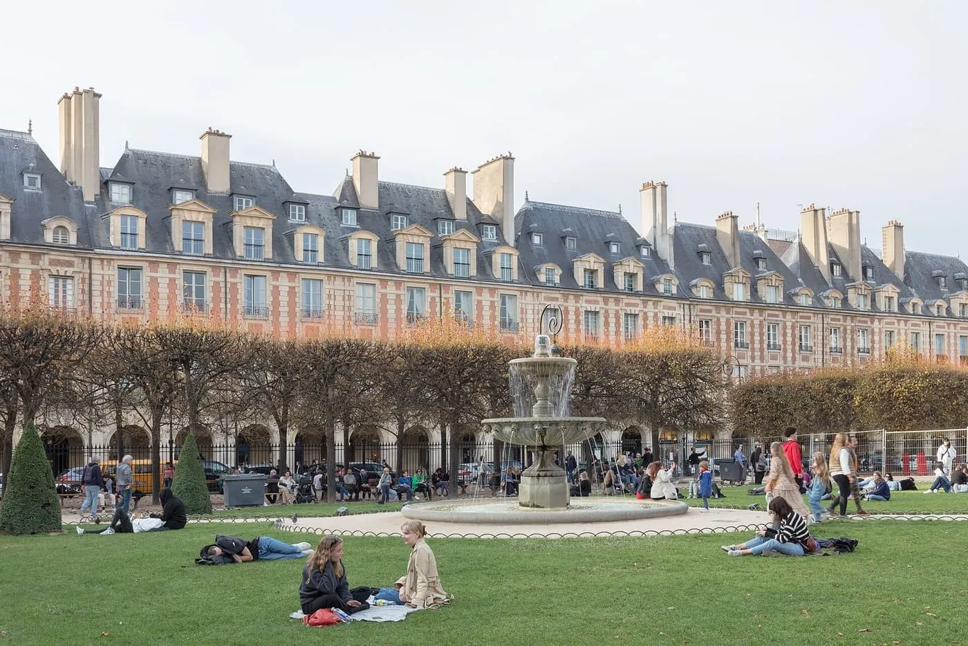 The brick-and-stone pavilions of the Place des Vosges behind a fountain and clipped lime trees, with people sitting on the lawn