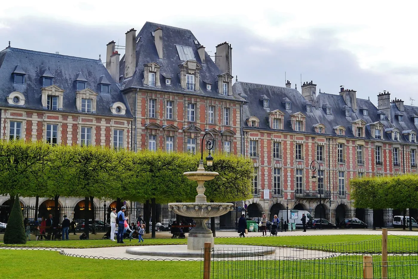 The red brick and stone pavilions of the Place des Vosges, with their slate roofs, behind a fountain and clipped lime trees in the square