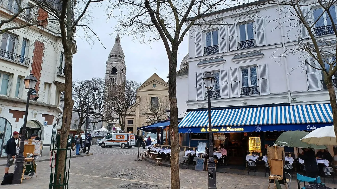 Café tables, easels and bare trees on Place du Tertre, with a tall pale bell tower rising behind the rooftops