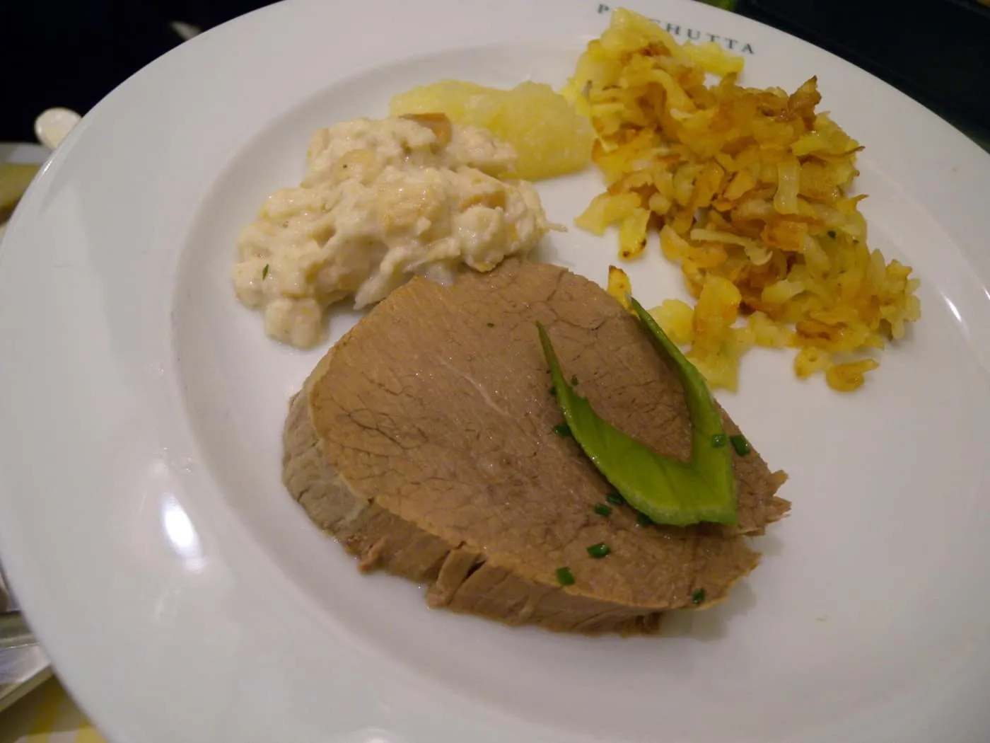 A slice of boiled beef with a chive-green garnish on a Plachutta plate, with a creamy side, apple sauce and rösti