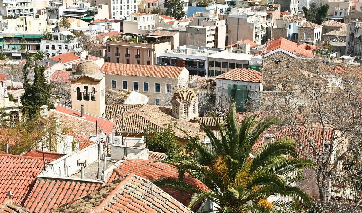 Terracotta rooftops of Plaka seen from above, with a bell tower and a small domed Byzantine church among the houses and a palm tree in the foreground