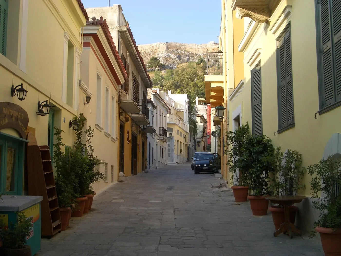 A quiet cobbled lane in Plaka between yellow neoclassical houses with shutters and potted plants, with the Acropolis rock closing the view